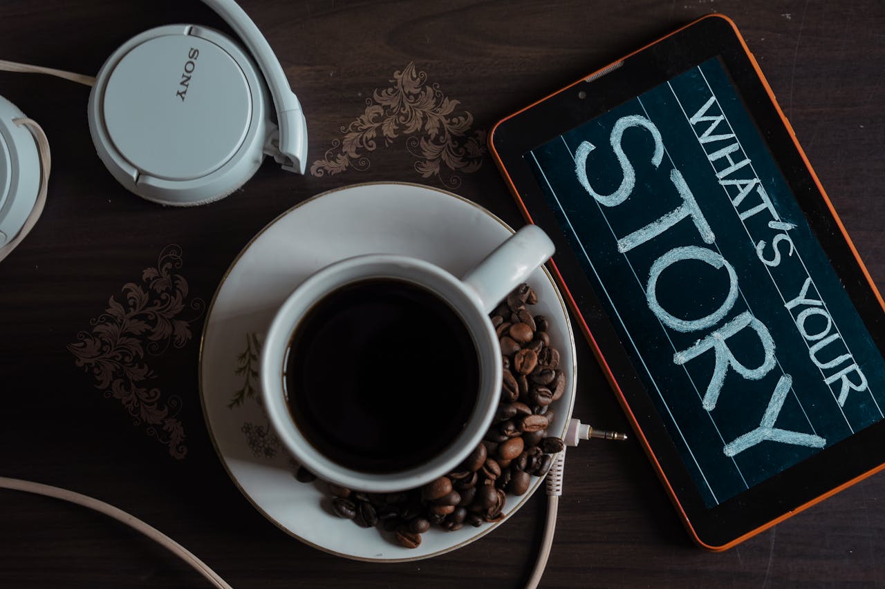 Flat lay of coffee cup, headphones, and tablet with Whats Your Story on screen.